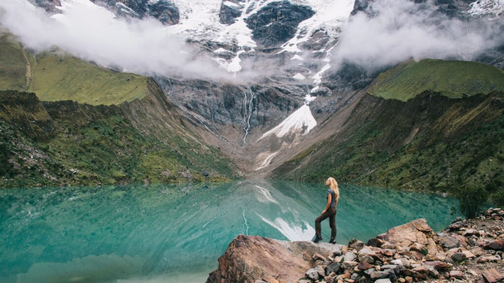 A traveler stands on a rocky edge overlooking the turquoise waters of Humantay Lake, surrounded by towering mountains and glacial peaks near Soraypampa.