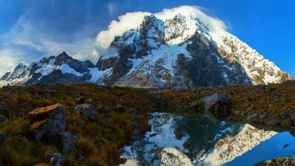 The stunning, snow-capped Salkantay Mountain is reflected in a small glacial pond, surrounded by rugged Andean terrain. A breathtaking view of one of Peru's most iconic peaks along the Salkantay Trek route - Inka Trail Peru.info.