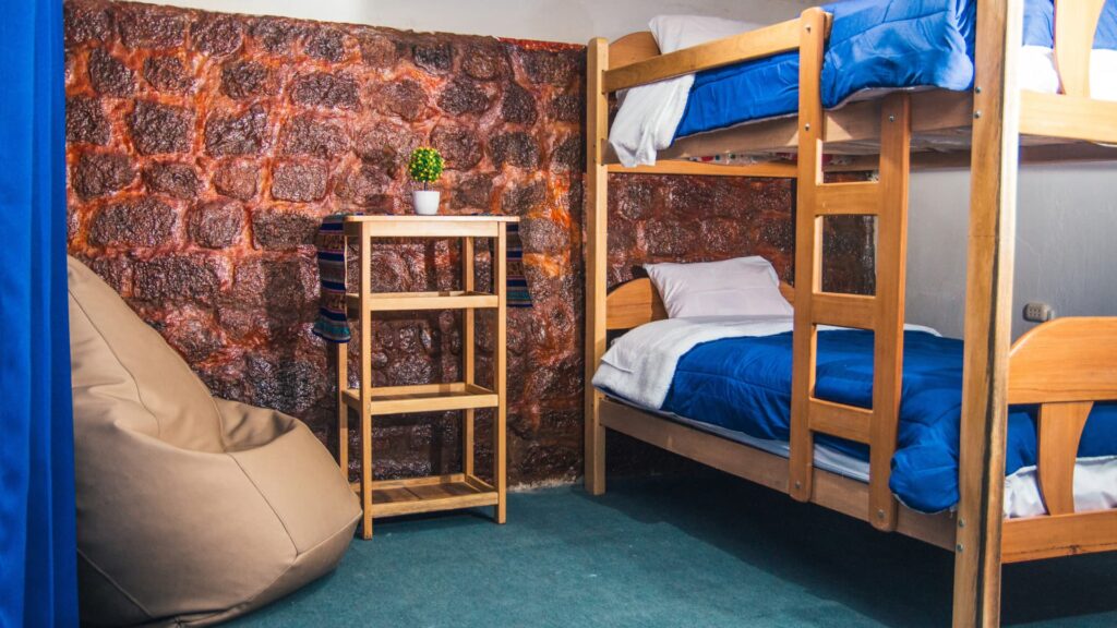 Interior of a shared room with wooden bunk beds and deep blue covers, offering scenic views of Cusco’s rooftops through a large window - IncaTrailPeru.info