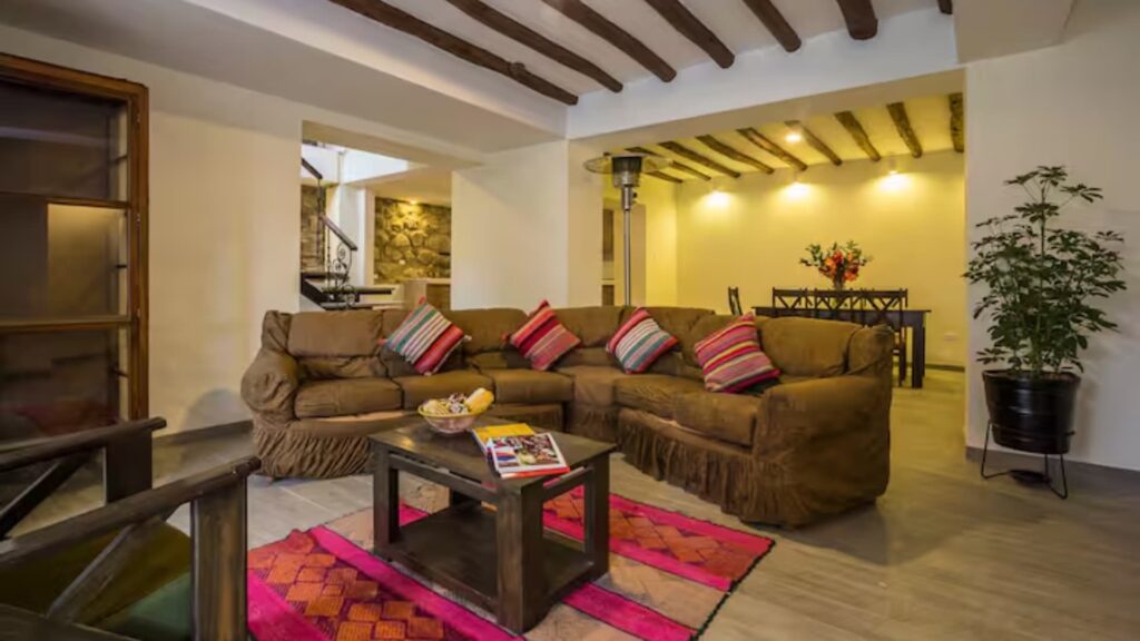 Spacious and cozy living room featuring a large brown sectional sofa, colorful Andean pillows, wooden beams, and a vibrant pink rug - IncaTrailPeru.info