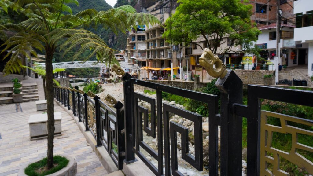 Decorative black and gold railings alongside a walkway in Aguas Calientes, with a backdrop of buildings, river, and lush mountains near Machu Picchu - IncaTrailPeru.info